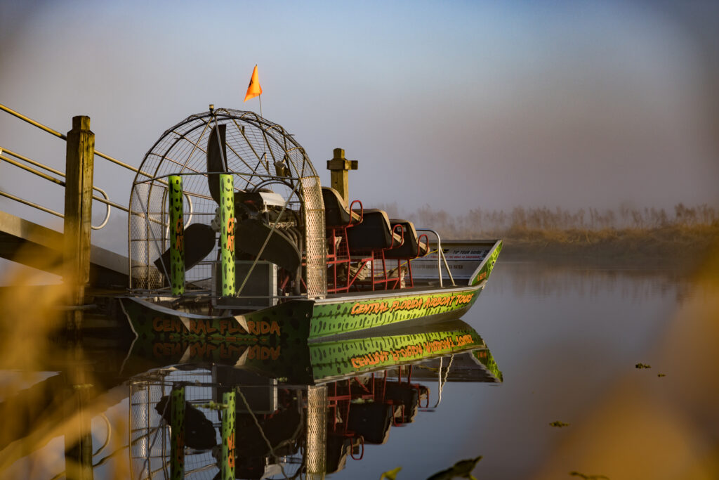 A green airboat with passengers glides over calm water at sunset.